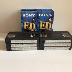 May include: Two blue Sony ED Max 6h T-120 VHS tape boxes positioned behind two stacks of three black VHS tapes. Each black tape features a white adhesive label with handwritten text detailing the recorded content and dates on the spine.