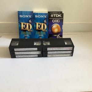 Peut inclure: Un ensemble de six cassettes VHS noires disposées en deux piles de trois, accompagnées de trois étuis en carton d'origine. Les étuis portent les marques Sony ED Max et TDK Q-HG, avec du texte indiquant 6 heures d'enregistrement par cassette.