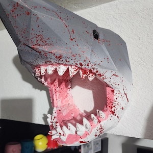 Puede incluir: Decoración de pared con forma de cabeza de tiburón geométrica gris con detalles de salpicaduras rojas. La boca del tiburón está abierta, revelando dientes rosas y blancos. Montada en una pared blanca.