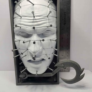 May include: A white, 3D printed, human face sculpture with a grid of nails protruding from the surface. The sculpture is in a gray box with a metal hook attached to the bottom.