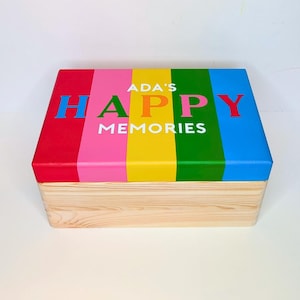 May include: A wooden box with a lid painted in a rainbow of colours. The lid is divided into vertical stripes of red, pink, yellow, green, and blue. The words "ADA'S HAPPY MEMORIES" are printed on the lid in white. The box is made of natural wood.