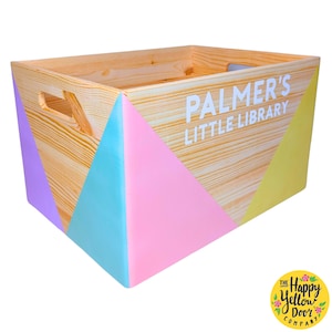 May include: Wooden storage box with a pastel pink, blue, and purple geometric design. The box is labeled "Palmer's Little Library" and has a handle on the side. The box is made by The Happy Yellow Door Company.