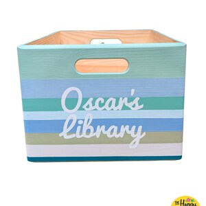 May include: A rectangular wooden storage crate featuring horizontal stripes in shades of teal, light blue, sage green, and white. The front panel displays the white text Oscar's library in a script font. The crate includes cutout handles on the sides.