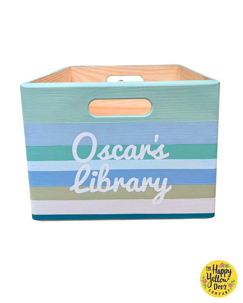 May include: A rectangular wooden storage crate featuring horizontal stripes in shades of teal, light blue, sage green, and white. The front panel displays the white text Oscar's library in a script font. The crate includes cutout handles on the sides.