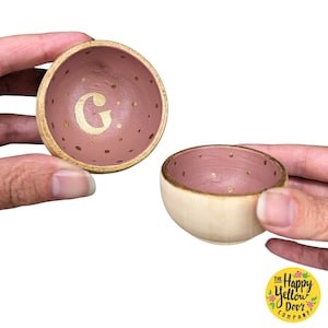 May include: Two small wooden bowls with a pink and gold speckled design. One bowl has the letter "G" painted in gold on the inside.
