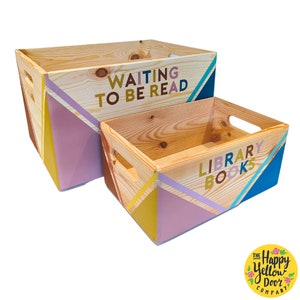 May include: Two rectangular wooden crates with cut-out handles, featuring natural wood grain and geometric painted corners in yellow, lavender, and blue. The larger crate displays the text WAITING TO BE READ, while the smaller crate displays LIBRARY BOOKS.
