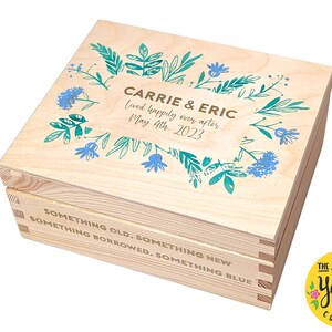 May include: A rectangular light-colored wooden box with a hinged lid. The lid features a blue and green floral wreath design surrounding the text CARRIE & ERIC, Lived happily ever after, May 4th, 2023. The front side displays the text SOMETHING OLD, SOMETHING NEW, SOMETHING BORROWED, SOMETHING BLUE.