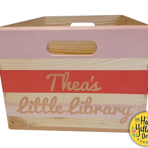 May include: A rectangular wooden storage crate featuring a light pink painted top section and a horizontal coral-colored stripe. The text "Thea's little library" is printed in a light color across the front. The crate includes integrated handles.