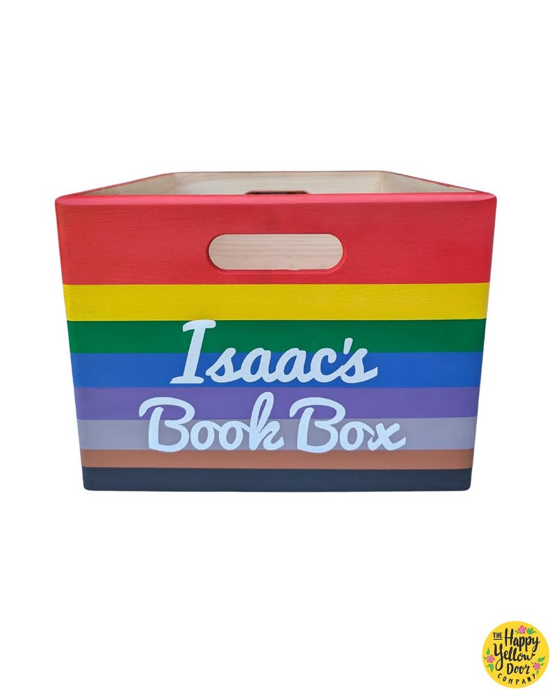 May include: A rectangular wooden storage crate featuring horizontal stripes in red, yellow, green, blue, purple, gray, brown, and black. The front panel displays the white text Isaac's Book Box and includes a cutout handle at the top center.