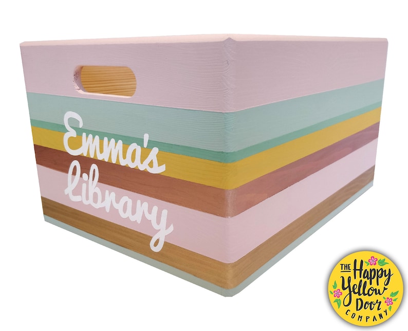 May include: A rectangular wooden storage crate featuring horizontal stripes in shades of light pink, sage green, mustard yellow, and brown. The front panel displays the white text "Emma's library" in a script font and includes a cutout handle.