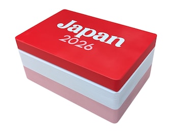 May include: A rectangular box with a stacked appearance featuring three distinct layers. The top layer is bright red with the white text Japan 2026 printed on it. The middle layer is white, and the bottom layer is a soft, muted pink color.