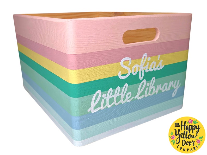 May include: A rectangular wooden storage crate featuring horizontal stripes in pastel pink, yellow, green, and blue. The front panel displays the white painted text "Sofia's little library" in a cursive script. The crate includes a cutout handle.