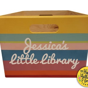 May include: A wooden storage crate featuring horizontal stripes in yellow, teal, light green, pink, blue, and orange. The front displays the white text "Jessica's little library" and includes a cutout handle at the top center of the box.