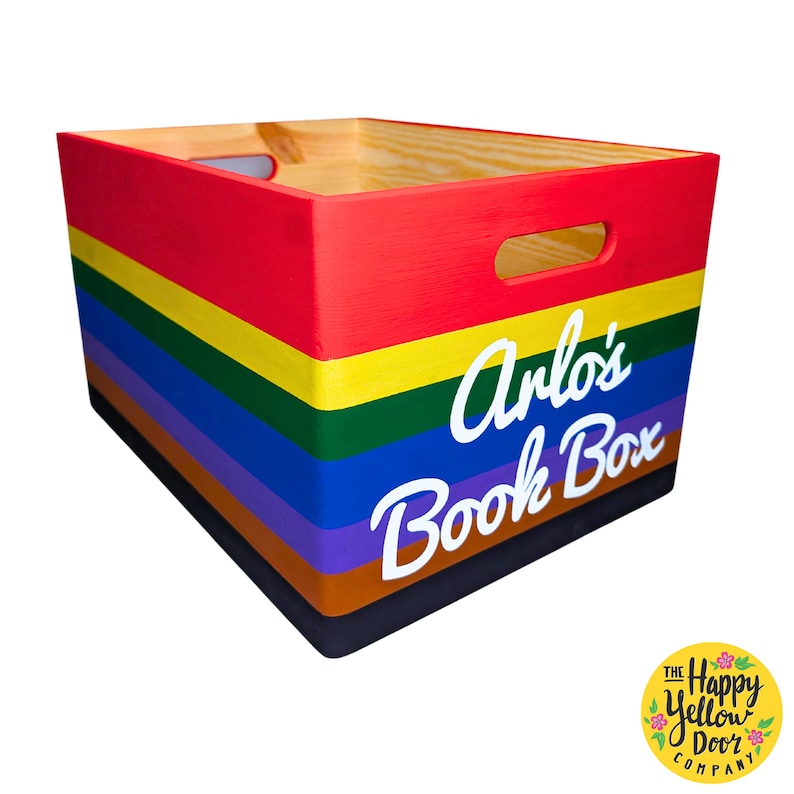 May include: A rectangular wooden storage crate featuring horizontal stripes in red, yellow, green, blue, purple, brown, and black. The front panel displays the white text Arlo's Book Box in a script font. The crate includes two oval handle cutouts.