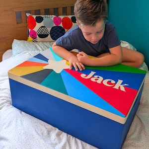May include: A blue toy chest with a colourful starburst design on the lid. The lid features the name "Jack" in white letters. The chest is rectangular with a light beige trim.