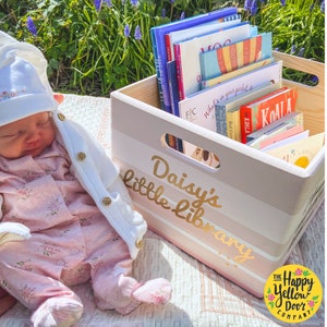 May include: A light-colored wooden storage crate with cut-out handles, featuring the gold text Daisy's little library, filled with a collection of colorful children's books. The crate is positioned next to a sleeping infant wearing a floral outfit.