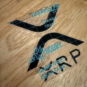 May include: Two clear vinyl decals with black and teal text and graphics. The decals read "STANDA TRAM" and "HOBBY HARD GRIP ISFER TAPE". The decals are on a light brown wooden surface.