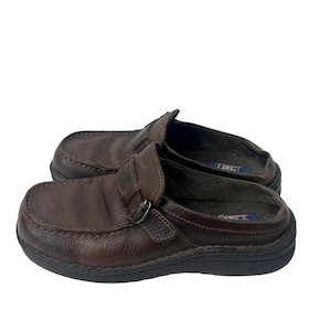 May include: A pair of brown leather slip-on shoes with a buckle detail. The shoes have a rounded toe and a black sole. The leather appears worn, suggesting they are pre-owned.