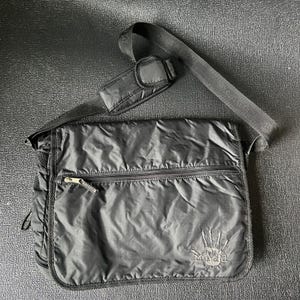 May include: Black messenger bag with a shoulder strap. The bag has a zippered front pocket and a handprint logo that says "Mud". The bag is made of a shiny, quilted material.