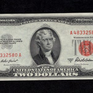 May include: A two-dollar bill featuring a portrait of Thomas Jefferson. The bill is primarily green and black, with red seals and serial numbers. The text "TWO DOLLARS" is printed multiple times around the edges.