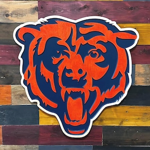 Op de afbeelding: Houten wandkunst met het logo van de Chicago Bears. Het logo is een berenkop in oranje en marineblauw met een witte omtrek. De achtergrond bestaat uit kleurrijke houten planken.