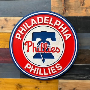 Op de afbeelding: Een rond houten wandbord met het logo van de Philadelphia Phillies. Het ontwerp bevat een blauw silhouet van de Liberty Bell, de rode tekst Phillies en de witte tekst Philadelphia Phillies, geplaatst tegen een felrode achtergrond met blauwe en witte randen.