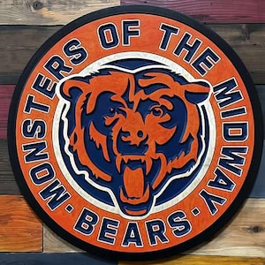 Op de afbeelding: Een rond houten bord met het Chicago Bears-logo, met de tekst "MONSTERS OF THE MIDWAY BEARS" in witte letters. Het berenlogo is oranje en marineblauw. Het bord staat tegen een veelkleurige houten plank achtergrond.