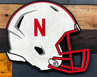 May include: Wooden cutout of a football helmet. The helmet is white with a red facemask and the letter "N" in red. The cutout is layered with black and gray accents.