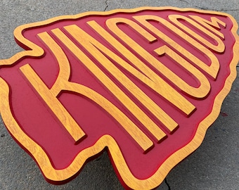 May include: A wooden sign in the shape of an arrowhead with the word "KINGDOM" carved into it. The sign is painted red with gold lettering.