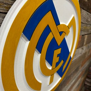 Pode incluir: Uma placa de madeira branca, azul e dourada com o bras&atilde;o do Real Madrid. O bras&atilde;o apresenta uma letra "M" branca com uma coroa azul no topo e uma letra "F" dourada dentro do "M".