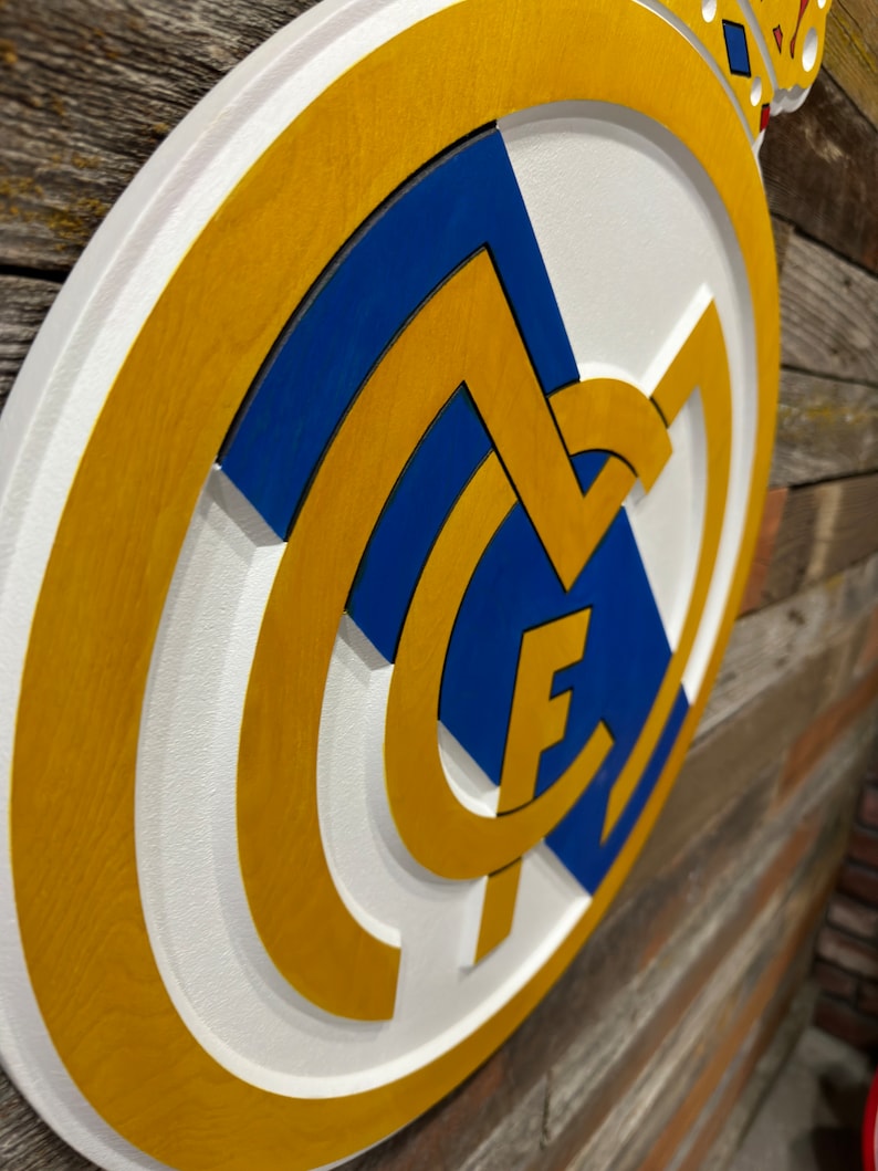 Pode incluir: Uma placa de madeira branca, azul e dourada com o bras&atilde;o do Real Madrid. O bras&atilde;o apresenta uma letra "M" branca com uma coroa azul no topo e uma letra "F" dourada dentro do "M".