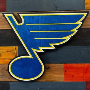 Op de afbeelding: Een houten bord met het logo van de St. Louis Blues. Het logo is een blauwe muzieknoot met een vleugelontwerp, omlijnd in zwart, wit en geel. Het bord is gemonteerd op een veelkleurige houten plank achtergrond.
