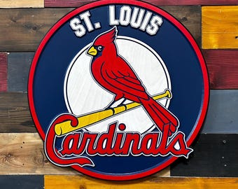 May include: A round wooden sign featuring the St. Louis Cardinals logo. The sign has a navy blue border with the words "ST. LOUIS" in white. A red cardinal bird is perched on a yellow baseball bat, with the word "Cardinals" in red.