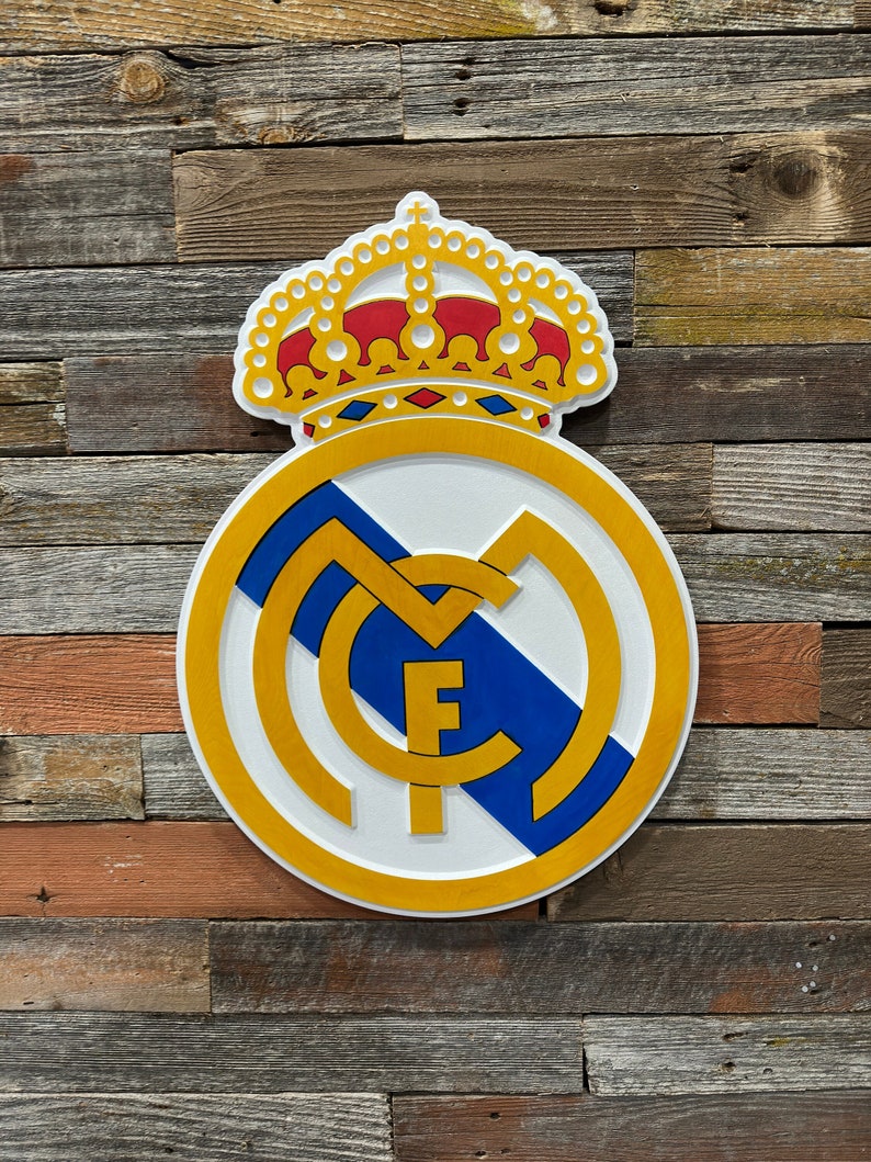 Pode incluir: Um logotipo de equipe de futebol branco e dourado com uma faixa azul e uma coroa dourada no topo. O logotipo est&aacute; em um fundo de madeira.