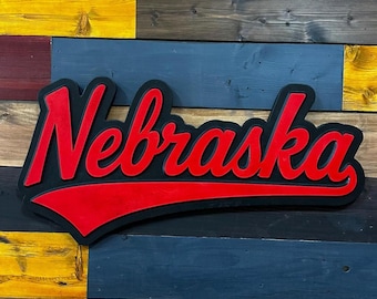 May include: A wooden sign with the word "Nebraska" in red, stylized script. The lettering is outlined in black and set against a black and red background. The sign is mounted on a multi-colored wooden plank wall.