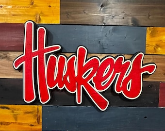 May include: A red and white wooden sign that reads "Huskers" in a cursive font. The sign has a black outline and is mounted on a multi-colored wooden plank background. The sign is a decorative item.