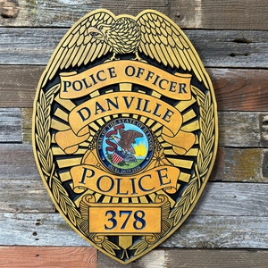 May include: A gold-colored police badge with a black eagle, the words "Police Officer Danville", and the seal of the State of Illinois. The badge also has the word "Police" and the number "378" on it.