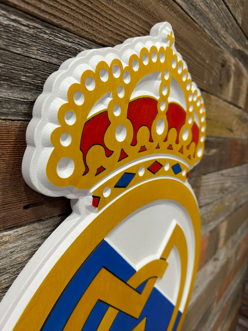 Pode incluir: Uma decora&ccedil;&atilde;o de parede de madeira branca, dourada e azul com o bras&atilde;o do Real Madrid. O bras&atilde;o inclui uma coroa com detalhes vermelhos e azuis.