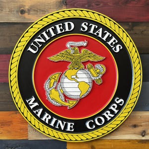 Op de afbeelding: Een rond houten bord met het embleem van het United States Marine Corps. Het bord heeft een rode kern met een gouden adelaar, wereldbol en anker. De tekst "UNITED STATES MARINE CORPS" is wit, met een gele touwrand.