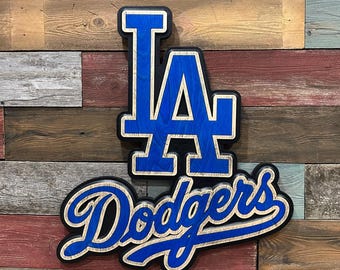 May include: A wooden sign with the Los Angeles Dodgers logo. The "LA" is in a blue and black font with a white outline. The word "Dodgers" is in a blue and black font with a white outline and a white swirl.