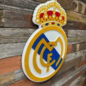 Pode incluir: Um sinal de madeira 3D branco, dourado e azul do bras&atilde;o do Real Madrid com uma coroa dourada no topo. O bras&atilde;o apresenta uma letra "M" branca com uma letra "F" dourada dentro, e uma faixa azul e branca.