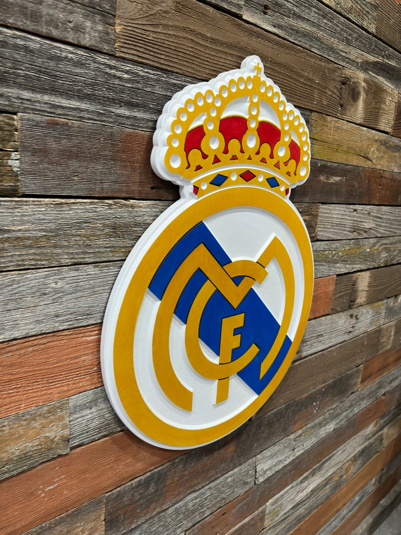 Pode incluir: Um sinal de madeira 3D branco, dourado e azul do bras&atilde;o do Real Madrid com uma coroa dourada no topo. O bras&atilde;o apresenta uma letra "M" branca com uma letra "F" dourada dentro, e uma faixa azul e branca.