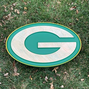 Op de afbeelding: Een ovaalvormig houten bord in groen en goud met een grote witte "G" in het midden. Het bord ligt op een bed van groen gras.