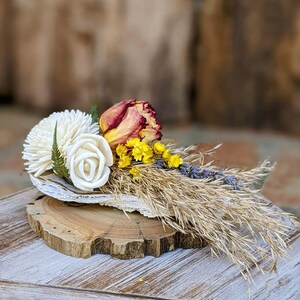 May include: A dried flower arrangement featuring white sola wood flowers, a dried rose, yellow billy buttons, and dried lavender sprigs. The arrangement is displayed on a wooden slice.