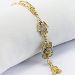 May include: Gold-toned charm featuring a Hamsa hand, a rectangular shape with an eye design, and a tassel. The charm is adorned with clear and turquoise-colored crystals. The chain is also gold-toned.