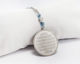 May include: A silver-colored round pendant with Arabic script, attached to a chain with blue beads and a decorative silver bead. The pendant is likely a religious or spiritual item, possibly a charm or amulet.