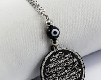 May include: A black pendant with Arabic script, suspended from a silver chain. The pendant features a circular design with intricate text and is adorned with a black bead with an eye symbol. The background is a white fabric.