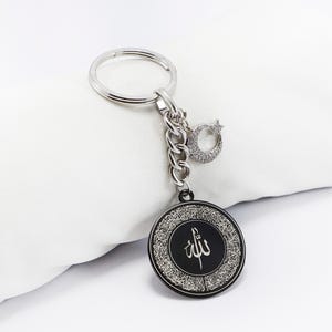 Op de afbeelding: Zilverkleurige sleutelhanger met een ronde zwarte hanger met Arabische kalligrafie en een decoratieve rand. De sleutelhanger bevat een ketting, een maanvormige bedel en een sleutelring.