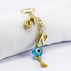 Puede incluir: Llavero dorado con las letras "S" y "N", un amuleto de ojo turco azul y una pequeña borla dorada. El llavero también presenta un anillo dorado y una pequeña perla. Las letras y los amuletos cuelgan de delicadas cadenas.