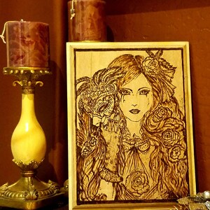 May include: A wood-burned image of a woman with long, flowing hair wearing a masquerade mask. The woman is holding the mask to her face with her right hand. The image is framed in a light wood frame.